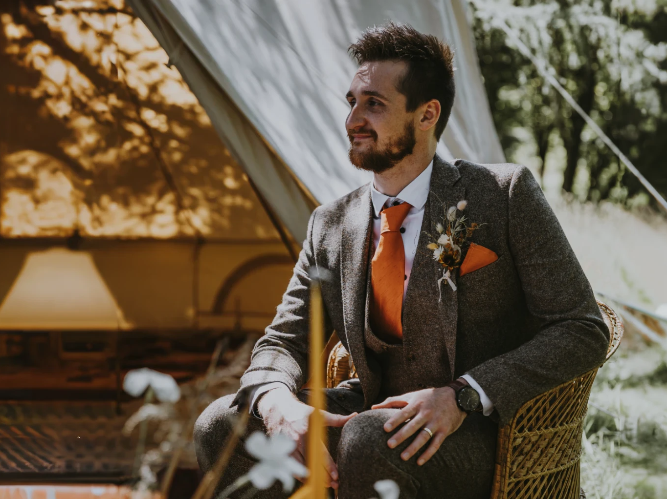 White groom seated outside in front of a teepee, wearing and orange tie