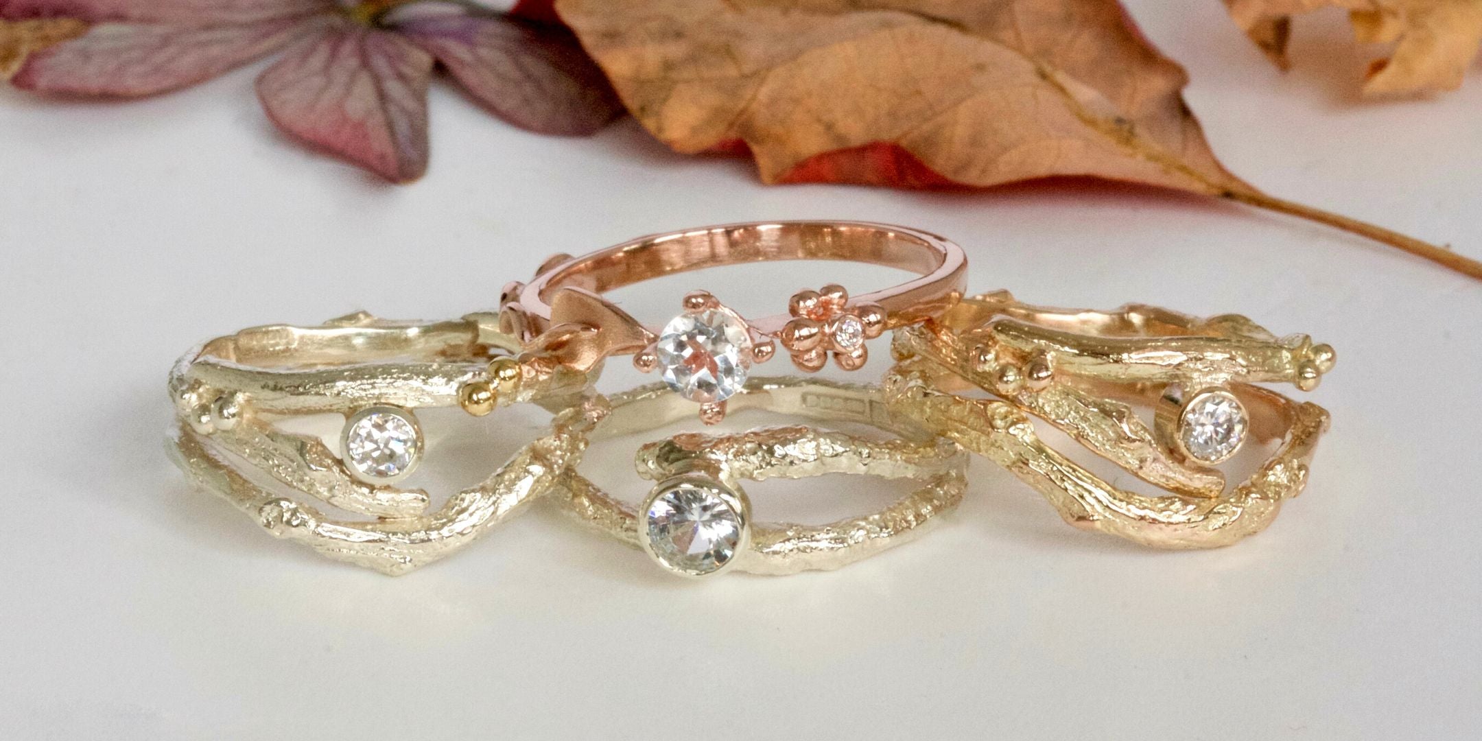 Three handmade gold rings and one rose gold ring on a white surface with autumnal leaves in the background