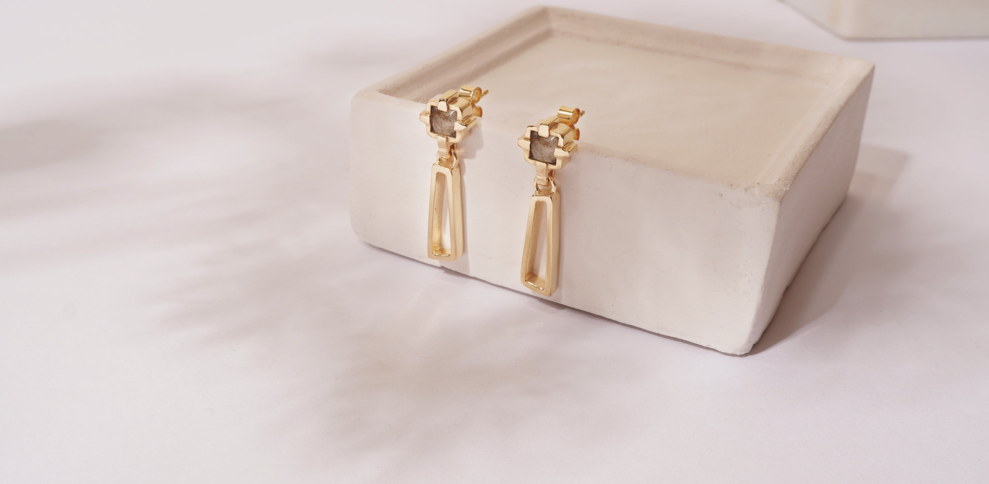 Two handmade gold drop earrings resting against a grey concrete block