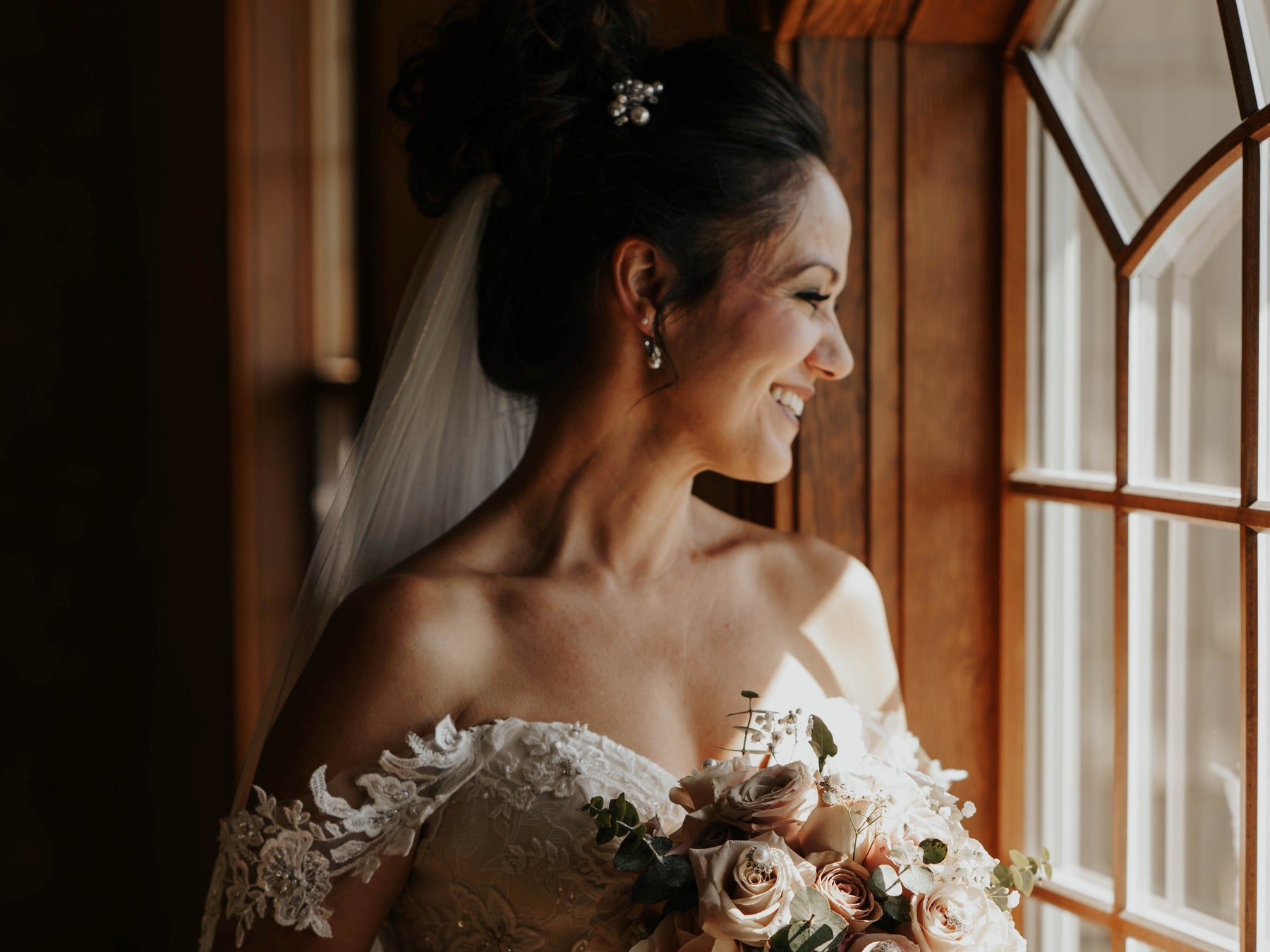 Bride standing next to a window holding a bouquet and wearing bespoke handmade jewellery